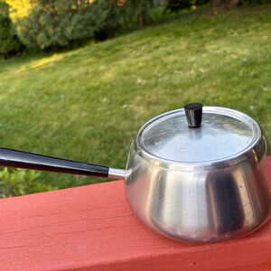 May include: A round, brushed stainless steel saucepan with a long, straight black handle and a matching circular lid. The lid features a small, cylindrical black knob centered on top. The pan has a smooth, tapered body and a polished metallic finish.
