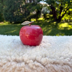 May include: A polished red stone apple figurine featuring a smooth, rounded body and a metallic gold-colored stem with a single leaf attached to the top. The object has a glossy finish that reflects light across its deep red surface.
