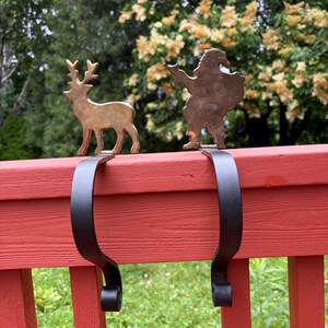 May include: Two metal stocking holders featuring a brass-toned reindeer and a brass-toned Santa Claus silhouette. Each figure is attached to a matte black curved metal hook designed to hang over a ledge, with a decorative scroll end at the base.