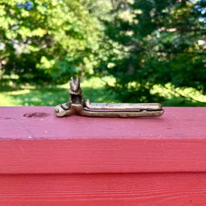 May include: A bronze-colored metal object, possibly a decorative latch or clasp, featuring a detailed design of a rabbit or hare. The object is resting on a red wooden surface, with a blurred green background of trees and foliage.