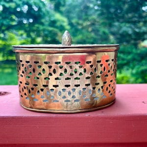 May include: A small, antique brass box with a lid and a decorative, openwork design. The box has a rounded shape and a small, acorn-shaped knob on top. The background is a blurred view of green foliage.