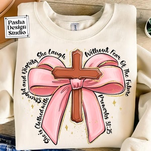 May include: A beige sweatshirt featuring a pink bow and a brown cross in the centre. The text "She is clothed with strength and dignity She laugh without fear of the future Proverbs 31:25" is written around the bow and cross.