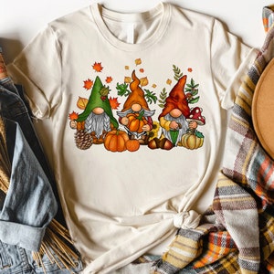 Puede incluir: Tres gnomos con sombreros de tem&aacute;tica oto&ntilde;al y sosteniendo calabazas. Los gnomos est&aacute;n rodeados de hojas de oto&ntilde;o, bellotas y un girasol.