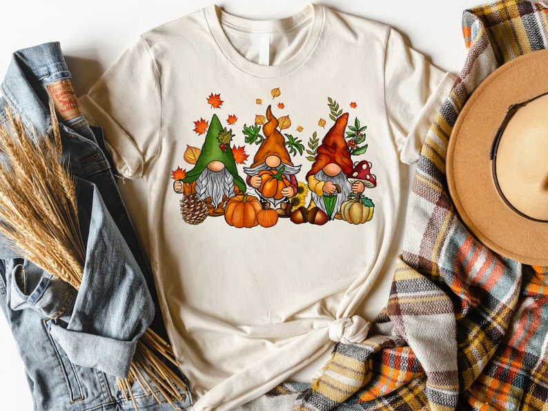 Puede incluir: Tres gnomos con sombreros de tem&aacute;tica oto&ntilde;al y sosteniendo calabazas. Los gnomos est&aacute;n rodeados de hojas de oto&ntilde;o, bellotas y un girasol.