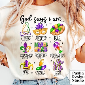 May include: A white t-shirt with a colorful Mardi Gras theme design. The design includes a crown, a fleur-de-lis, a king cake, a mask, and a lobster. The text "God says I am..." is at the top of the design, followed by a list of words and bible verses.