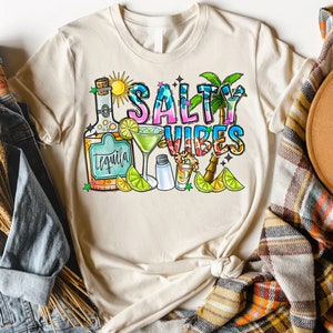 May include: A beige t-shirt with the text "Salty Vibes" in colorful letters. The design includes a bottle of tequila, a margarita glass, a salt shaker, a shot glass, limes, and a palm tree.