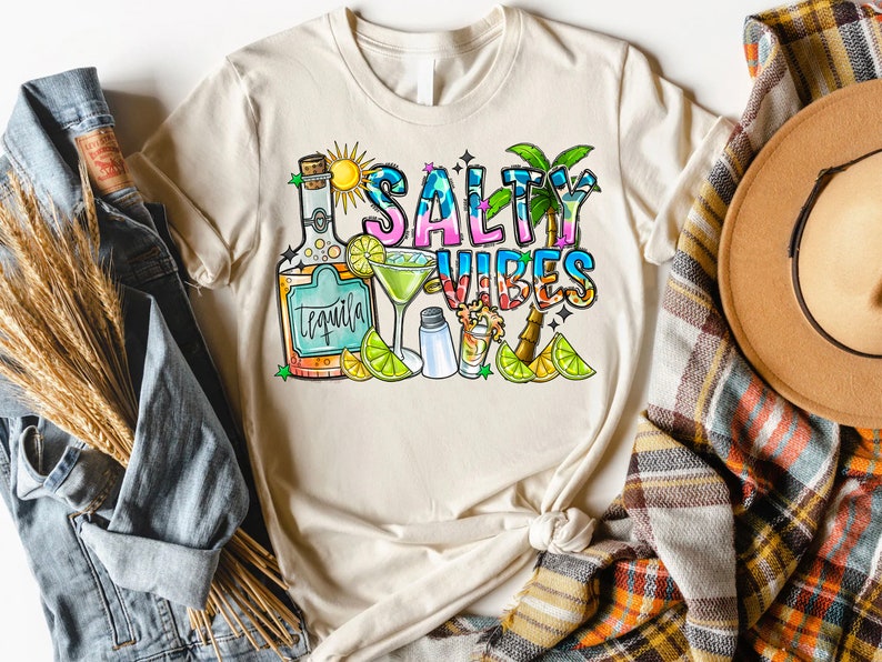 May include: A beige t-shirt with the text "Salty Vibes" in colorful letters. The design includes a bottle of tequila, a margarita glass, a salt shaker, a shot glass, limes, and a palm tree.