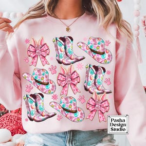 Peut inclure: Sweat-shirt rose avec un motif de P&acirc;ques color&eacute; comprenant des n&oelig;uds de lapin, des chapeaux de cow-boy et des bottes. Le motif comprend des couleurs pastel et des &oelig;ufs. Le texte "Pasha Design Studio" est dans le coin inf&eacute;rieur droit.