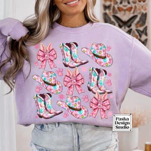 Peut inclure: Un sweat-shirt lavande avec un motif de P&acirc;ques color&eacute; comprenant des n&oelig;uds roses, des bottes de cow-boy et des chapeaux avec des &oelig;ufs de P&acirc;ques. Le motif est r&eacute;p&eacute;t&eacute; sur tout le sweat-shirt. Le texte "Pasha Design Studio" est visible dans le coin inf&eacute;rieur droit.