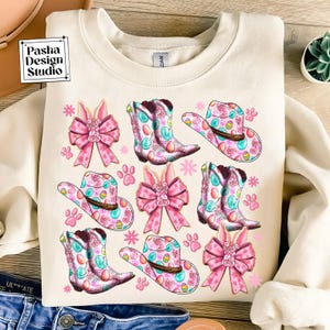 Peut inclure: Un sweat-shirt cr&egrave;me avec un motif r&eacute;p&eacute;t&eacute; de n&oelig;uds roses, de chapeaux de cow-boy et de bottes d&eacute;cor&eacute;es d'&oelig;ufs de P&acirc;ques pastel. Les chapeaux et les bottes ont un motif floral rose et bleu.