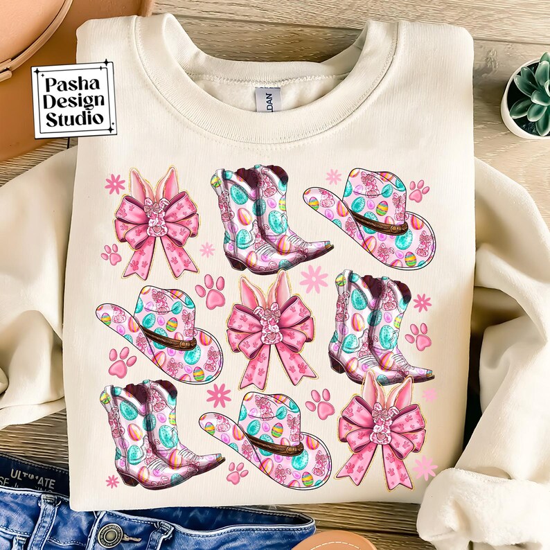 Peut inclure: Un sweat-shirt cr&egrave;me avec un motif r&eacute;p&eacute;t&eacute; de n&oelig;uds roses, de chapeaux de cow-boy et de bottes d&eacute;cor&eacute;es d'&oelig;ufs de P&acirc;ques pastel. Les chapeaux et les bottes ont un motif floral rose et bleu.