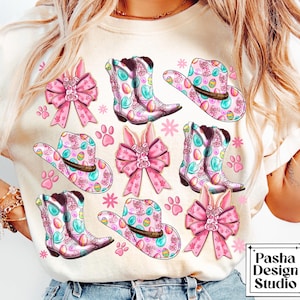 Peut inclure: Un t-shirt blanc avec un motif de P&acirc;ques rose et bleu. Le motif repr&eacute;sente des bottes de cow-boy, des chapeaux, des n&oelig;uds et des &oelig;ufs. Le texte "Pasha Design Studio" est visible dans le coin inf&eacute;rieur droit.