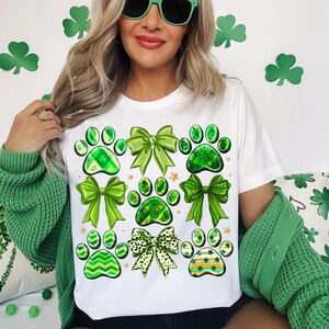 May include: White t-shirt with a green shamrock paw print design. The design features multiple paw prints with green shamrock patterns and green bows.