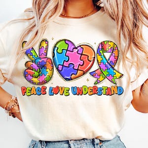 Może przedstawiać: Biała koszulka z symbolem pokoju, sercem i wstążką wykonaną z puzzli w różnych kolorach. Pod symbolami nadrukowany jest tekst "Peace Love Understand".
