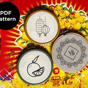 May include: Three embroidery hoops with different designs. The first hoop has a design of two Chinese lanterns. The second hoop has a design of a Chinese mooncake with the Chinese character for "fortune" inside. The third hoop has a design of an orange with a slice of orange next to it.