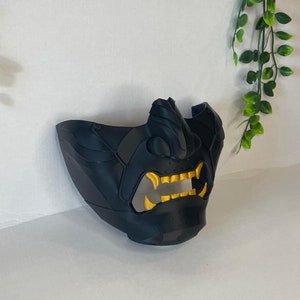 Op de afbeelding: Zwart en gouden samurai-masker met een woeste uitdrukking. Het masker is gemaakt van plastic en heeft een getextureerd oppervlak.