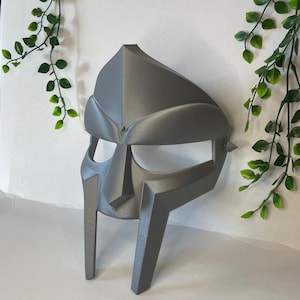 Puede incluir: Una máscara gris impresa en 3D con un diseño distintivo, que se asemeja a un casco con una parte superior puntiaguda y dos grandes cuernos curvos.