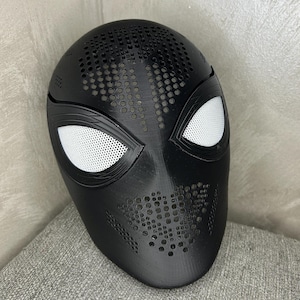 Può includere: Una maschera integrale nera con una superficie testurizzata caratterizzata da numerosi piccoli fori di ventilazione circolari sulla fronte e sulla bocca. La maschera presenta due grandi aperture oculari a mandorla coperte da una rete bianca.