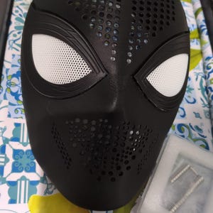 Peut inclure: Un masque intégral noir mat présentant une texture perforée sur le front et la zone de la bouche, avec de grandes ouvertures oculaires en forme d'amande recouvertes de lentilles en maille blanche. Un petit boîtier en plastique contient deux broches.