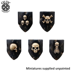 May include: Five black shield-shaped miniature plaques featuring raised gold-colored skull and bone motifs. The designs include three small skulls, a skull with crossbones, a side profile of a skull, a single skull, and a skull with a rib cage.