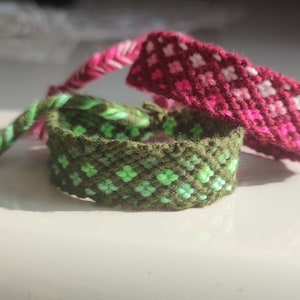 May include: Three friendship bracelets with a diamond pattern. One bracelet is green and white, one is pink and white, and one is dark green and light green.