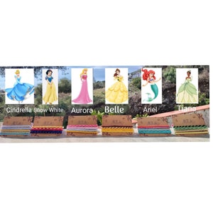 May include: Six colorful bracelets with different patterns, each featuring a Disney princess character: Cinderella, Snow White, Aurora, Belle, Ariel, and Tiana. The bracelets are displayed on a wooden surface.