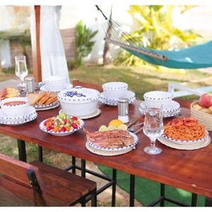 Peut inclure: Une table dressée avec des bols et des assiettes en céramique blanche avec une bordure bleue. La table est dressée avec une variété d'aliments, dont des pâtes, de la salade et du poisson. Il y a un hamac en arrière-plan.