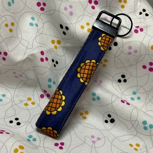 May include: A navy blue wrist strap key fob featuring a sunflower design in yellow and orange. It has a black metal clasp and key ring. The background is a white fabric with a pattern of grey lines and colourful dots.