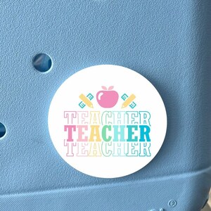 May include: A white round sticker with a red apple, two yellow pencils, and the text "TEACHER TEACHER TEACHER" in rainbow colors. The sticker is attached to a light blue container with the word "BOGG" printed on it.