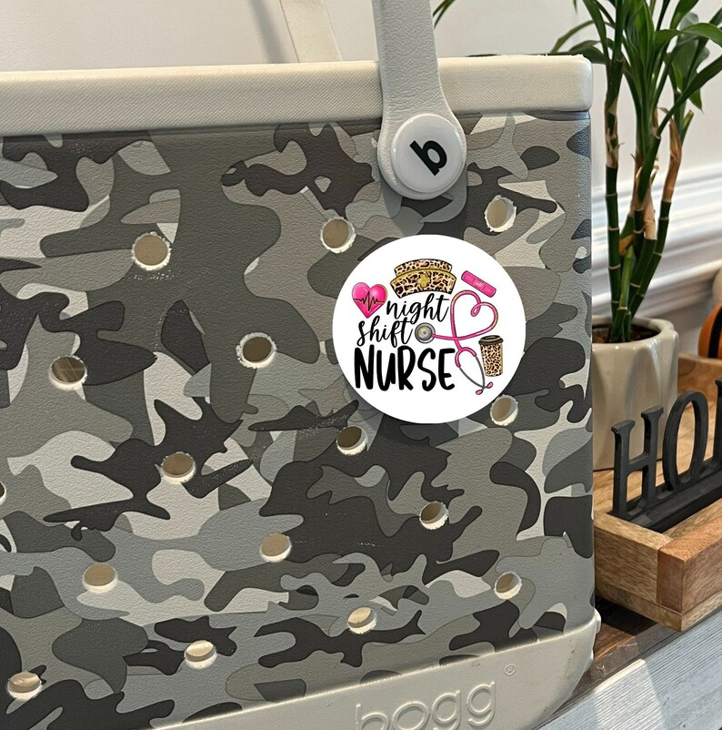 May include: A white round sticker with a pink heart, a stethoscope, a coffee cup, and the text "night shift nurse" in black. The sticker is on a gray and white camouflage patterned tote bag.
