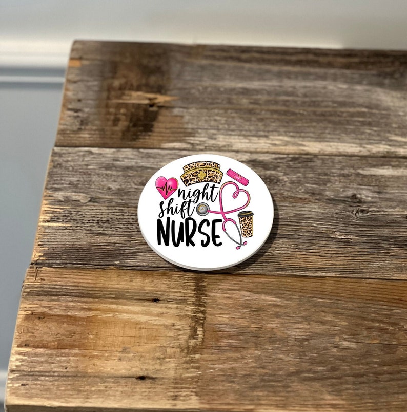 May include: White round coaster with a pink heart, stethoscope, coffee cup, and a leopard print nurse's hat. The text reads "night shift NURSE".