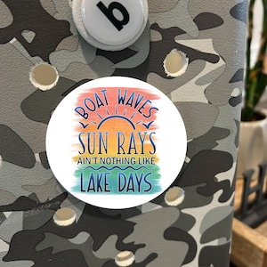 May include: A white, round decorative charm with the text "Boat Waves Sun Rays Ain't Nothing Like Lake Days" in blue, orange, and green. The charm is set against a camouflage pattern background.