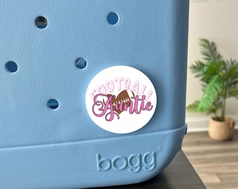 May include: A white round sticker with the text "Football Auntie" in pink lettering. A brown football with white laces is inside a pink heart. The sticker is attached to a light blue cooler with the word "bogg" printed on the front.