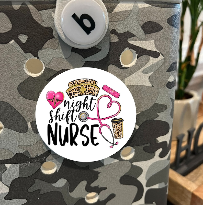May include: A white round sticker with a pink heart, a stethoscope, a coffee cup, and a leopard print nurse's hat. The text on the sticker reads "night shift NURSE".