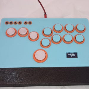 May include: A light blue arcade controller featuring orange-rimmed buttons with white and light blue caps. The device includes a small rectangular screen displaying a white graphic, a black base, and a red braided cable extending from the top edge.