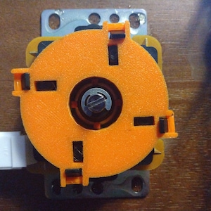 Puede incluir: Joystick de arcade naranja con un centro negro y cuatro botones direccionales negros. El joystick está montado en una placa de metal plateado.