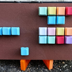 May include: A brown foam board with a variety of colorful keycaps arranged in different patterns. The keycaps are in shades of blue, green, yellow, orange, pink, and red. The board is sitting on a pair of orange stands.