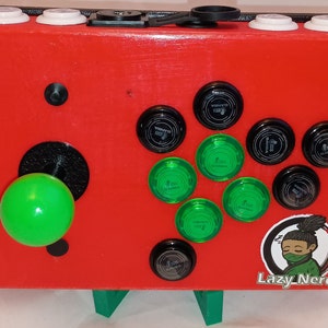 Op de afbeelding: Een rode arcadekast met een groene joystick en zwarte en groene knoppen. De kast heeft een zwart-wit logo met de tekst "Lazy Nerds".