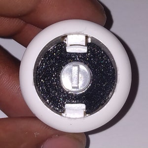 Puede incluir: Un recipiente redondo de pl&aacute;stico blanco con un centro negro y un bot&oacute;n plateado. El recipiente tiene una superficie texturizada y una peque&ntilde;a abertura en el lateral.