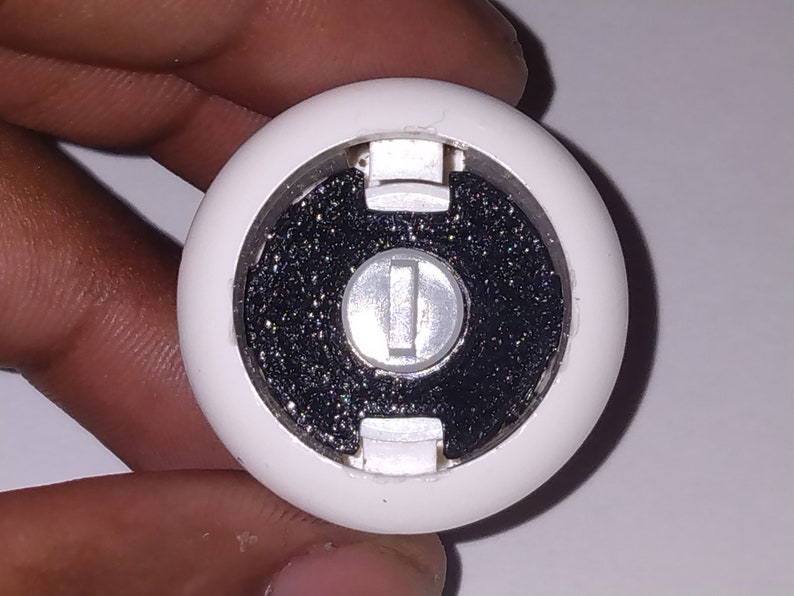 Puede incluir: Un recipiente redondo de pl&aacute;stico blanco con un centro negro y un bot&oacute;n plateado. El recipiente tiene una superficie texturizada y una peque&ntilde;a abertura en el lateral.