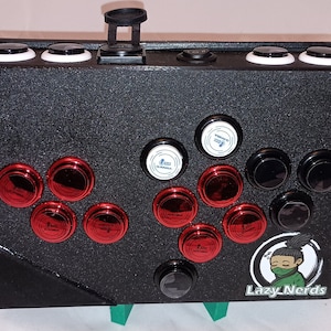 Puede incluir: Una palanca de arcade negra con botones rojos y negros. La palanca tiene un logotipo en blanco y negro que dice "Lazy Nerds".
