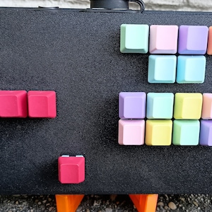 Könnte beinhalten: Eine Sammlung bunter Keycaps für eine mechanische Tastatur. Die Keycaps sind in einem Raster angeordnet und umfassen Farbtöne von Rosa, Lila, Blau, Gelb und Orange.
