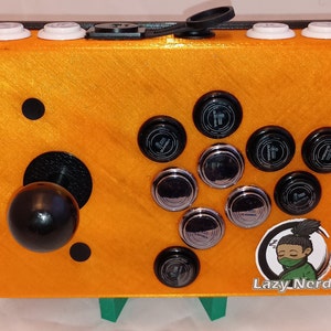 以下が含まれることがあります： オレンジ色のアーケードキャビネットに、黒いジョイスティックとボタンが付いています。キャビネットには、漫画のキャラクターと「Lazy Nerds」という文字が書かれた白黒のステッカーが貼られています。