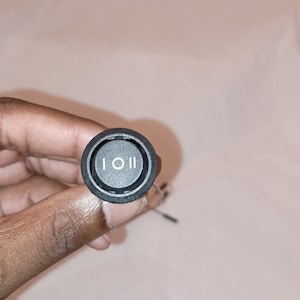 May include: A black round toggle switch with a white symbol of a circle with two vertical lines on either side. The switch is held in a hand.