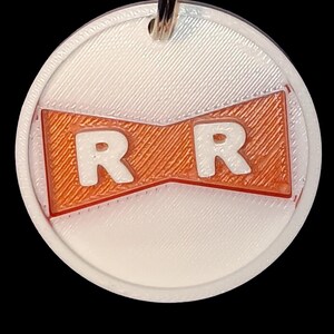 May include: A white and orange circular pendant with the letters "R R" in white. The pendant has a silver-colored metal loop at the top for attaching to a keychain or necklace. The design is a stylized logo.