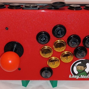 以下が含まれることがあります： ジョイスティック、ボタン、Lazy Nerdsのロゴが付いた赤いアーケードキャビネット。キャビネットは黒と金の色合いで、漫画のキャラクターで飾られています。