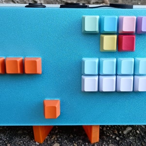 Puede incluir: Un teclado azul y naranja con una variedad de teclas de colores. El teclado tiene un fondo azul claro y está hecho de plástico. Las teclas están dispuestas en una cuadrícula y están etiquetadas con letras y números. El teclado está sentado en una mesa.