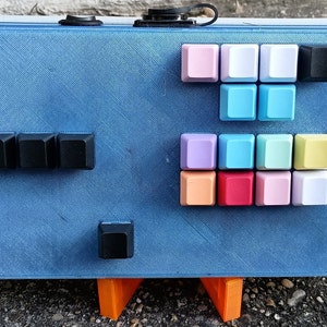 May include: A blue box with a variety of colored keycaps, including black, white, pink, light blue, lavender, peach, red, and yellow. The box is sitting on two orange stands.