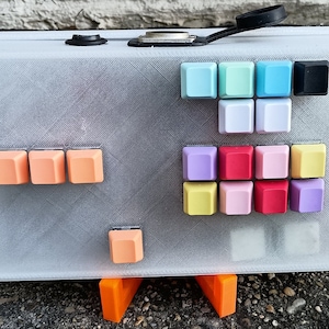 May include: A white plastic case with a variety of colorful keycaps. The case has a textured surface and is sitting on a stand. The keycaps are arranged in rows and columns, and they are all different colors. The keycaps are made of plastic and have a smooth surface.