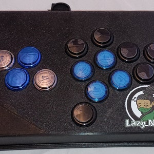 May include: A black arcade stick with blue and silver buttons. The stick has a black and white sticker with a cartoon character and the text "Lazy Nerds".